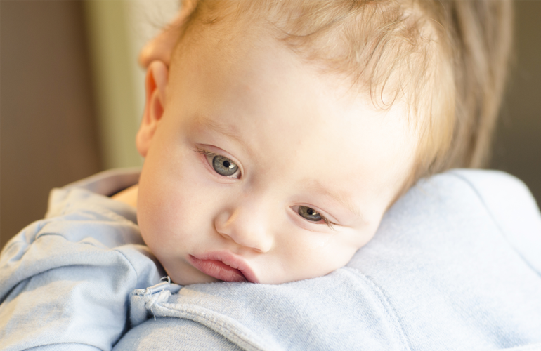 Ein müdes Baby mit blondem Haar und hellen Augen liegt mit dem Kopf auf der Schulter eines Erwachsenen. Das Baby trägt ein hellblaues Oberteil und wirkt erschöpft oder schläfrig. Der Hintergrund ist unscharf, wodurch der Fokus auf dem Gesicht des Babys liegt.